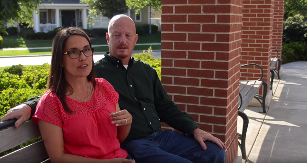 A man and a woman sitting side by side on a park bench. the woman, wearing glasses and a pink blouse, is talking, and the man in a green shirt attentively listens. they are surrounded by lush greenery and a brick structure.
