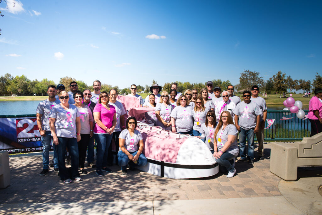 A group of people gathered around a large pink ribbon statue in a park, wearing pink and white shirts, showing support for breast cancer awareness.