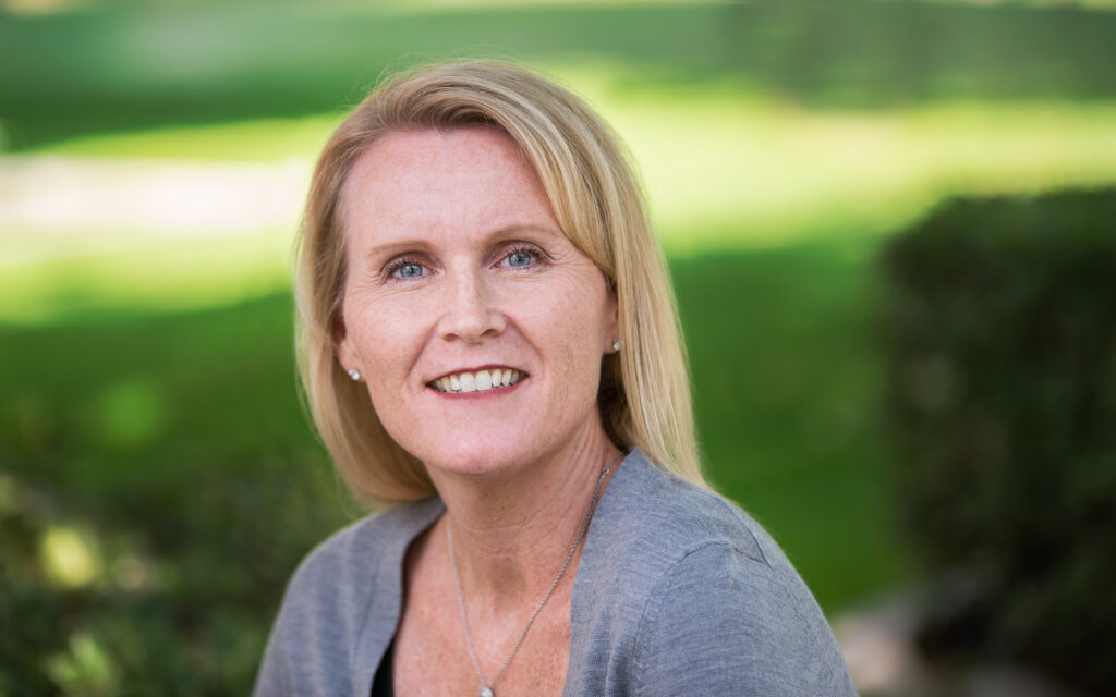 A smiling middle-aged woman with shoulder-length blonde hair, wearing a grey top, set against a soft-focus background of greenery and sunlight.