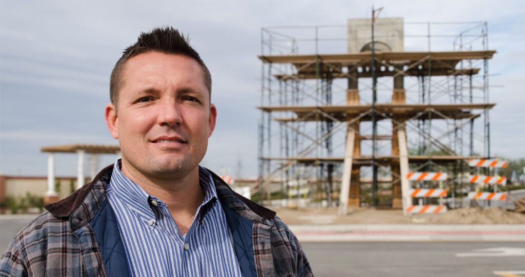 A man in a plaid shirt stands smiling in front of a construction site with scaffolding and a concrete structure in the background.