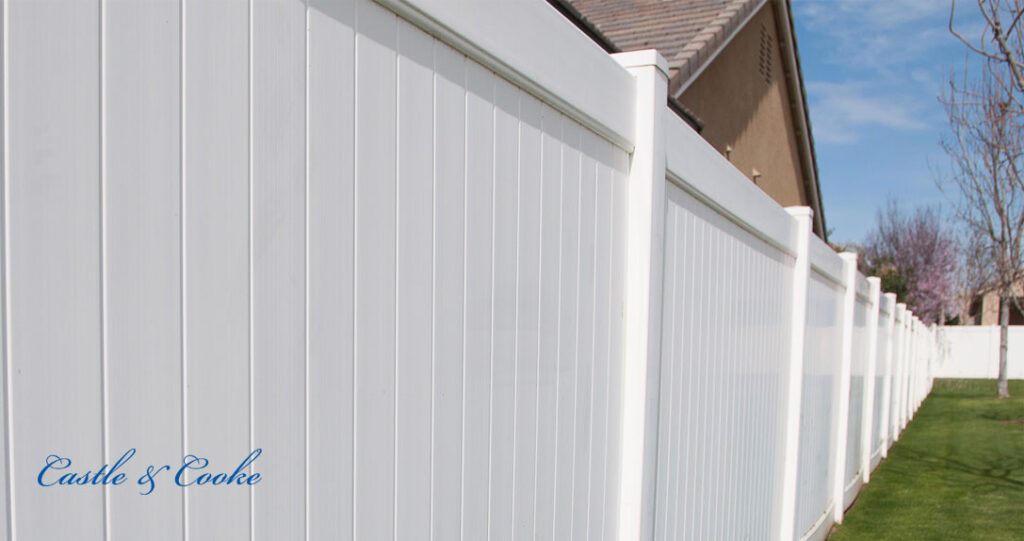 A long, white vinyl fence extends alongside a grassy area, with a clear blue sky above and a partial view of a house in the background. the text "castle & cooke" is visible at the bottom.