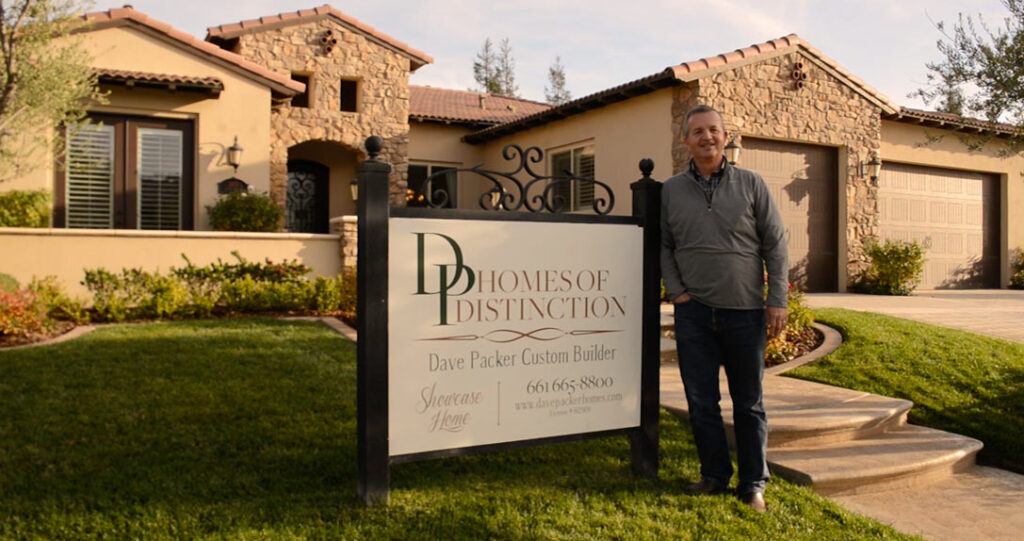 A man stands beside a sign reading "dp homes of distinction, dave packer custom builder" in front of a suburban home with a well-manicured lawn and a clear sky.