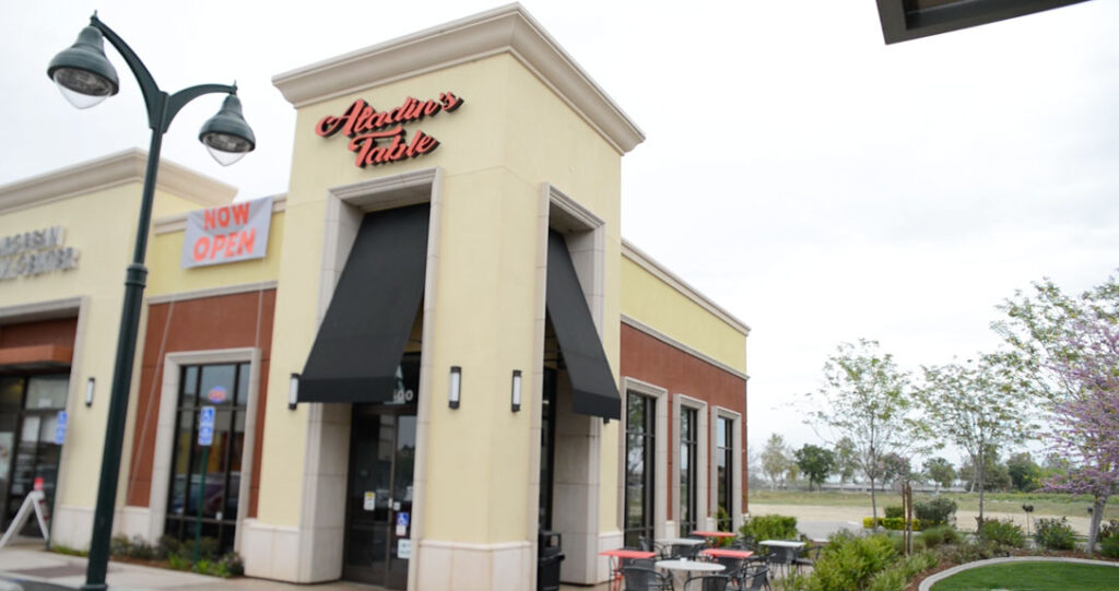 Exterior view of hector's taste restaurant with black awnings, located in a shopping center under a cloudy sky, featuring a few outdoor tables and a lush green lawn on the side.
