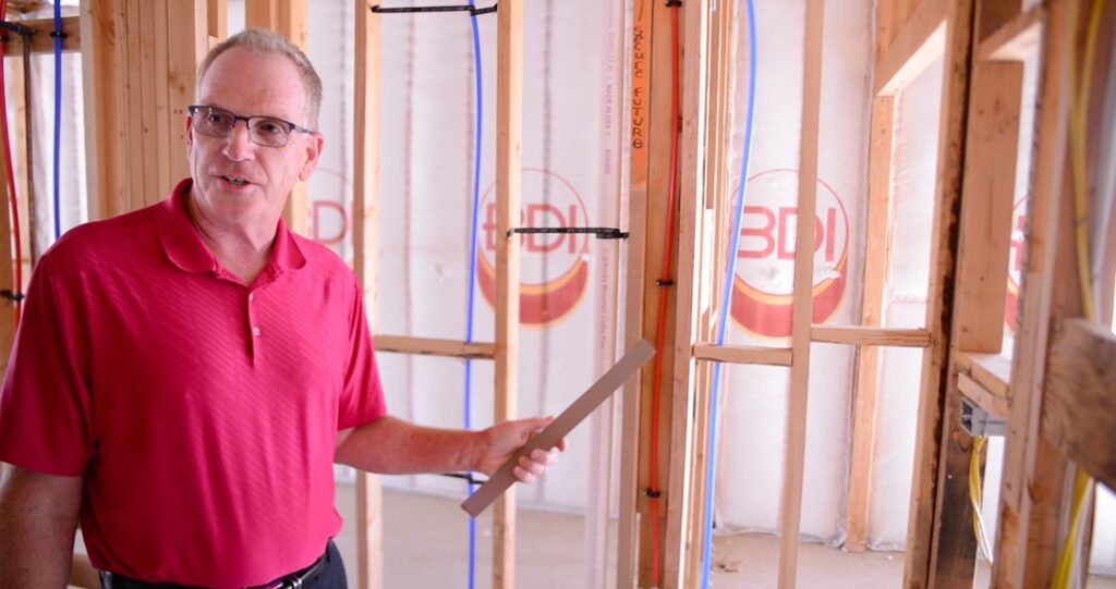 A man in a red polo shirt stands inside a construction site, holding a tablet and gesturing towards the unfinished walls with visible plumbing.