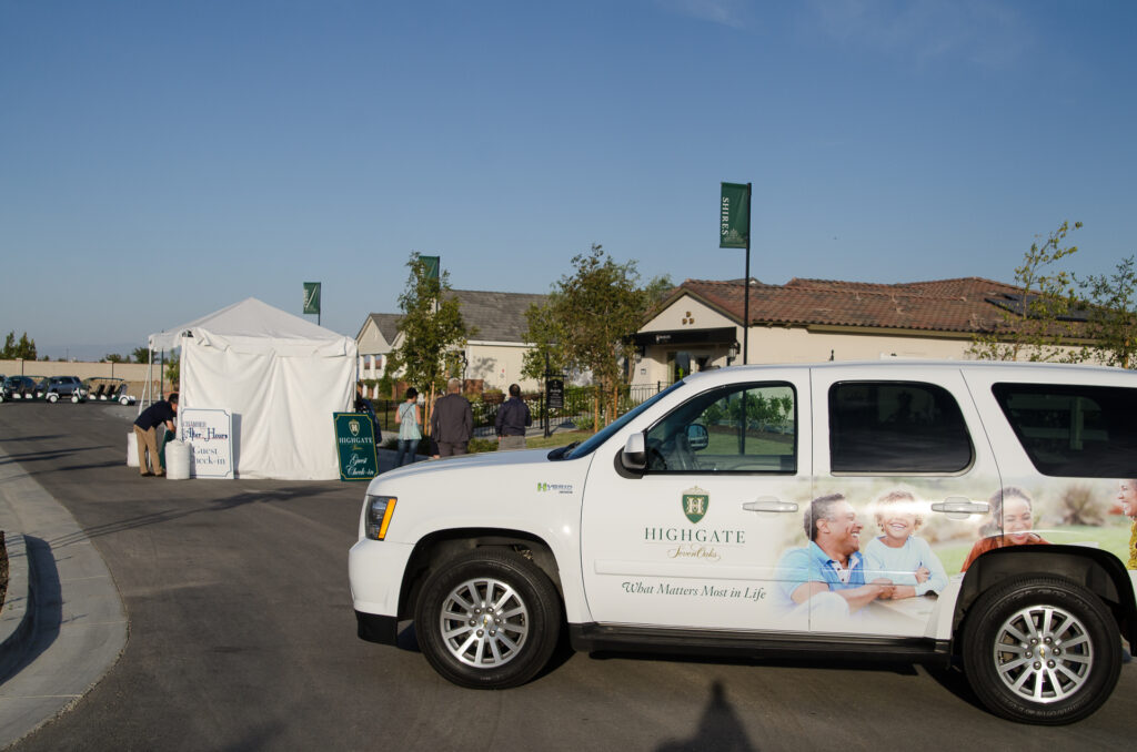 White suv with "highgate senior living" branding parked on a sunny street during an event, with tents and people in the background.