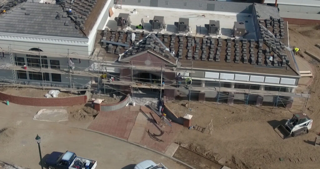 Aerial view of a construction site showing workers on scaffolding around a building with a classic façade, surrounded by construction equipment and partially landscaped areas.