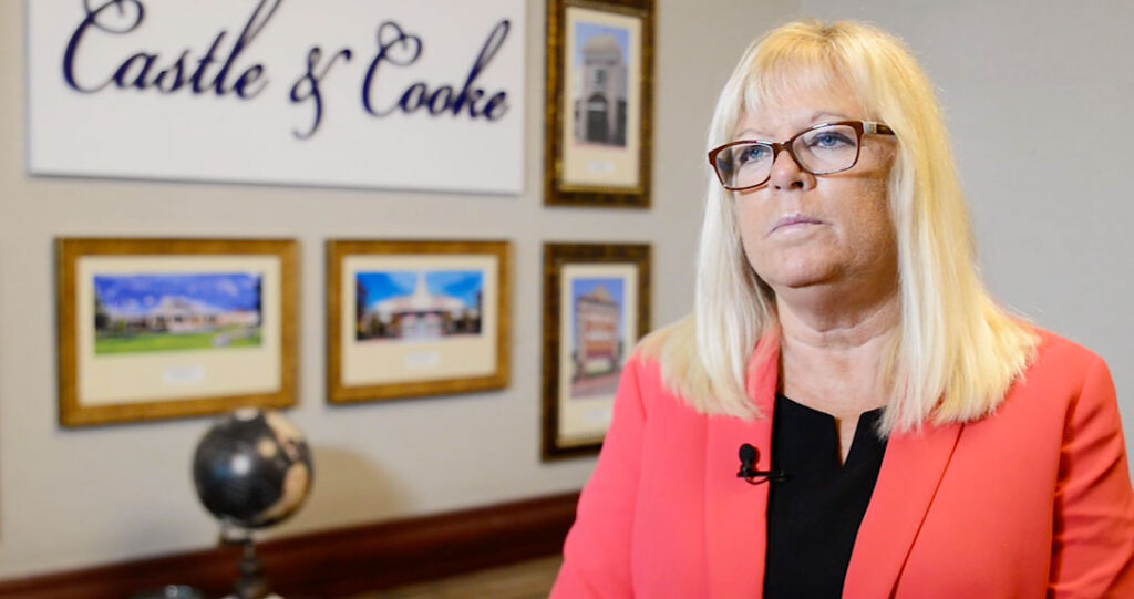 A woman in a red blazer and glasses stands in an office, with the "castle & cooke" sign and framed pictures of homes on the wall behind her. a globe is visible to her right.
