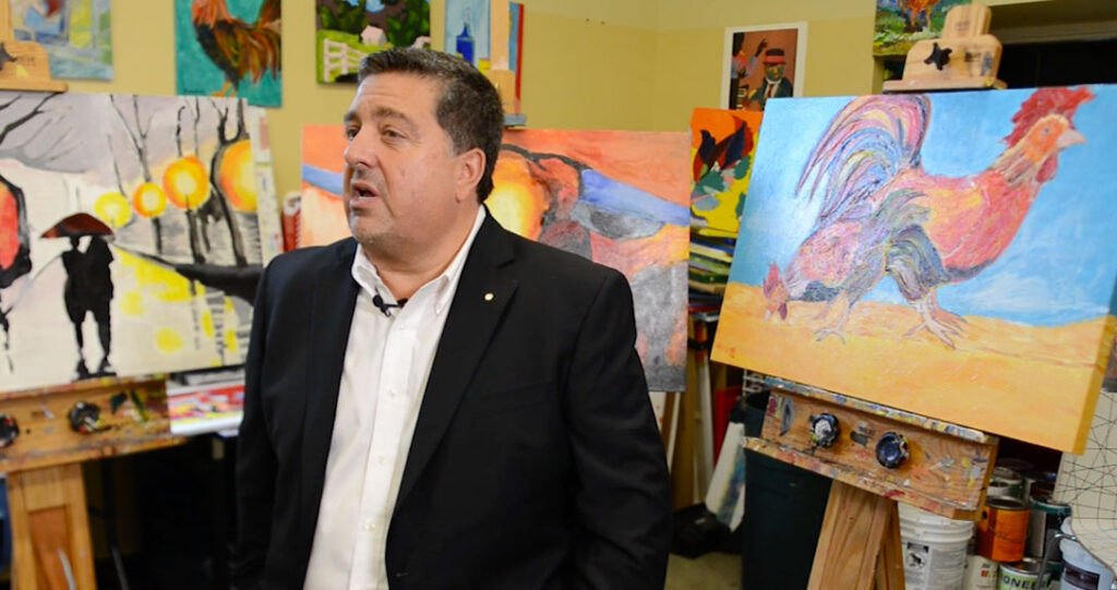 A man in a dark blazer talks passionately in an art studio filled with colorful paintings, including a prominent one of a rooster on an easel beside him.