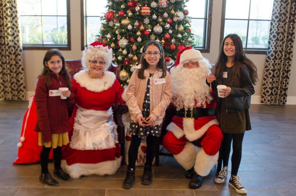 Four children pose with santa claus and mrs. claus by a large christmas tree. they wear seasonal attire and hold cups, smiling for the photo in a festively decorated room.