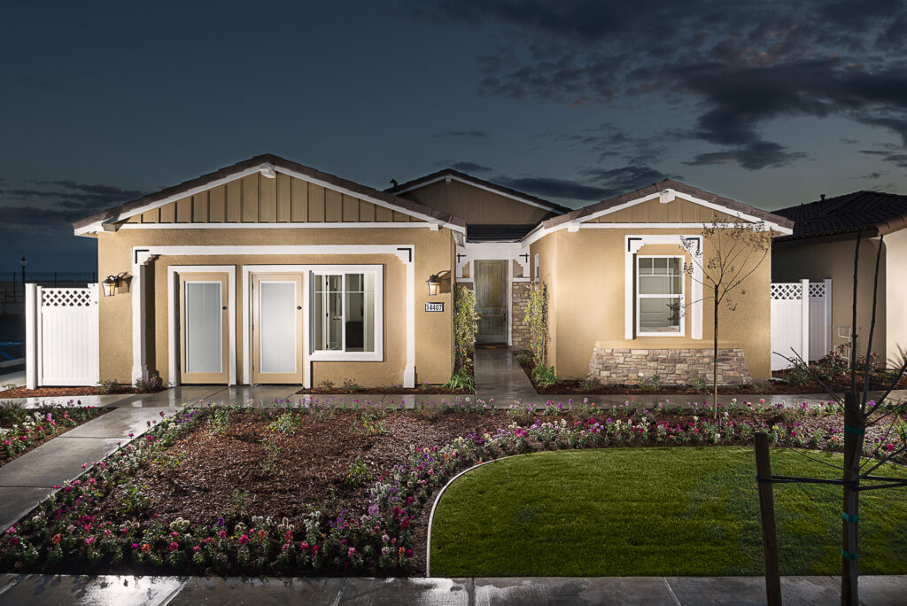 A modern single-story house with beige walls and a dark roof at dusk. the landscaping includes well-manicured grass and various flowers. a clear blue pool can be seen on the left.