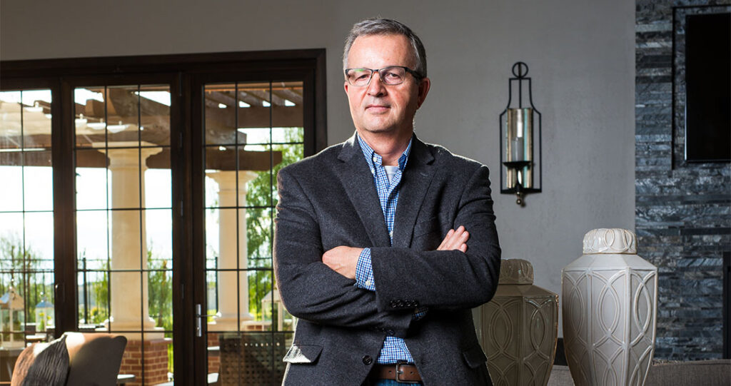 A confident man in glasses, wearing a blazer and a checkered shirt, stands with crossed arms in a stylish office with glass doors and elegant decor.