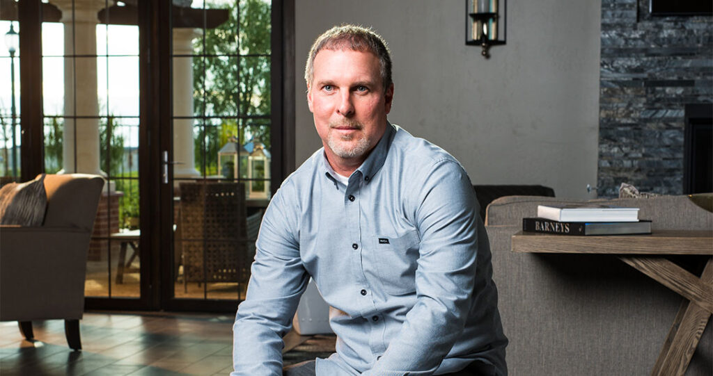 A man in a blue shirt sitting on the arm of a sofa in a stylish room with stone walls and large windows. books are on a coffee table beside him.