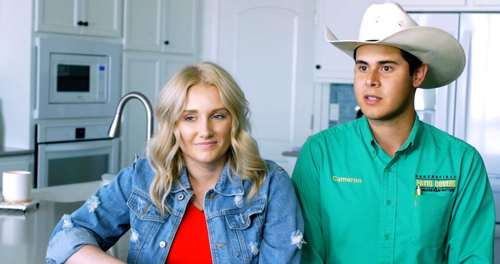 A woman with blonde hair and a man in a cowboy hat and green shirt standing in a kitchen, both looking to the side with a thoughtful expression.