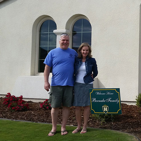 A man and woman smiling, standing next to a “welcome home” sign with the name “bernabe family” on a lawn in front of a house with large windows. A man and woman smiling, standing next to a "welcome home" sign with the name "bernabe family" on a lawn in front of a house with large windows.