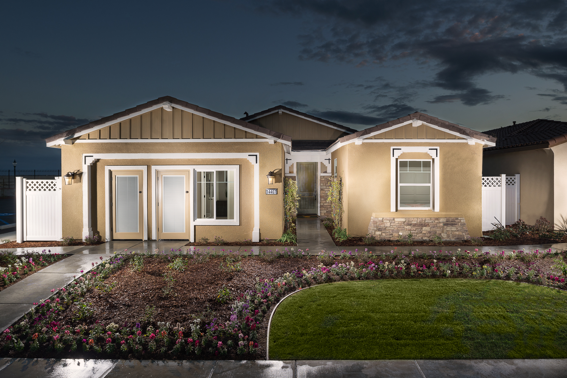 A new single-story house with beige stucco walls and a brown roof at twilight. the home features a landscaped, curved front yard with vibrant flowers, a trimmed lawn, and a paved path leading to the white front door.