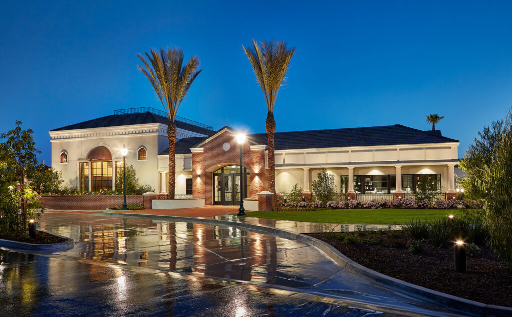 Luxurious mediterranean villa at twilight with wet driveway, bright interior lights, palms, and landscaped garden under a clear, dusky sky.