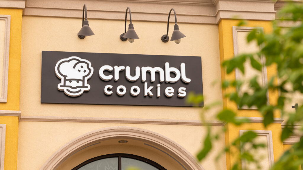 Exterior view of a "crumbl cookies" store sign mounted on a yellow wall above an arched doorway, lit by three gooseneck lamps.