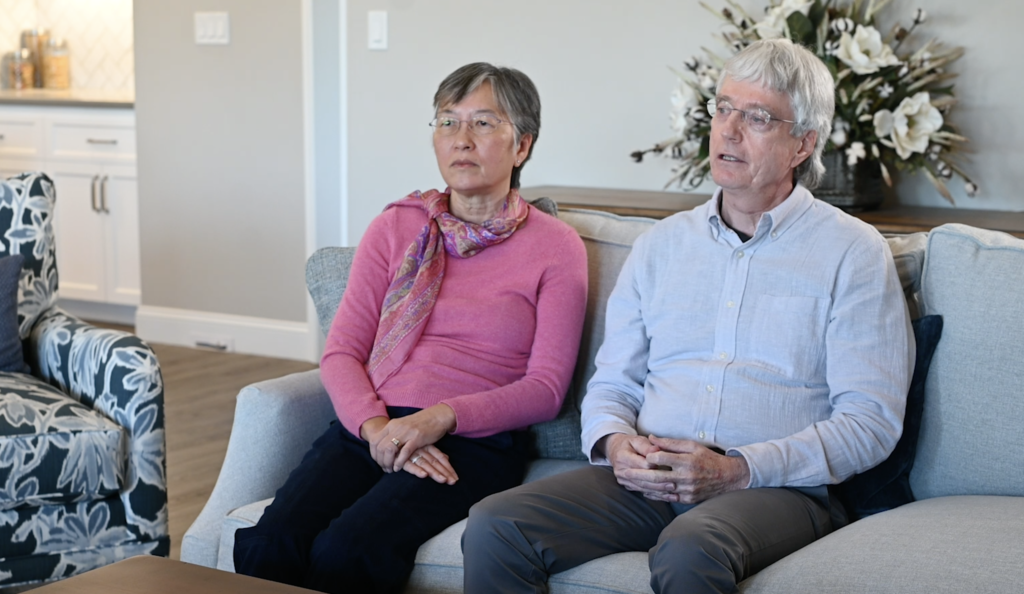 An elderly couple sits together on a sofa in a well-lit living room, looking slightly to their left with attentive expressions. the woman is wearing a pink sweater and a scarf, and the man a light blue shirt.
