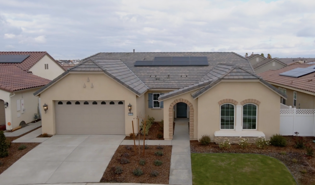 Aerial view of a single-story suburban home with a gray shingle roof featuring solar panels, arched windows, and a landscaped front yard.