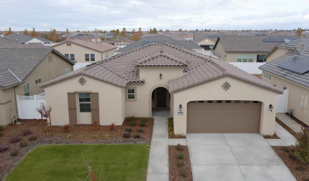 Aerial view of a single-story suburban home with a tan stucco exterior, terracotta roof tiles, a grassy front yard, and neighboring houses in similar styles.