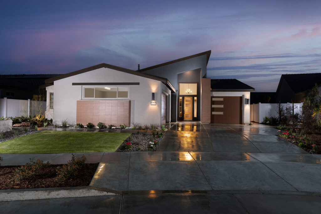 Modern single-story house at twilight with illuminated exterior lights, featuring a landscaped garden, and a concrete driveway leading to the front entrance.