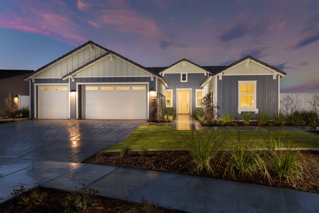 A modern single-story house at dusk, featuring a white and gray exterior, a double garage, and illuminated entrance, set against a vibrant sunset sky.