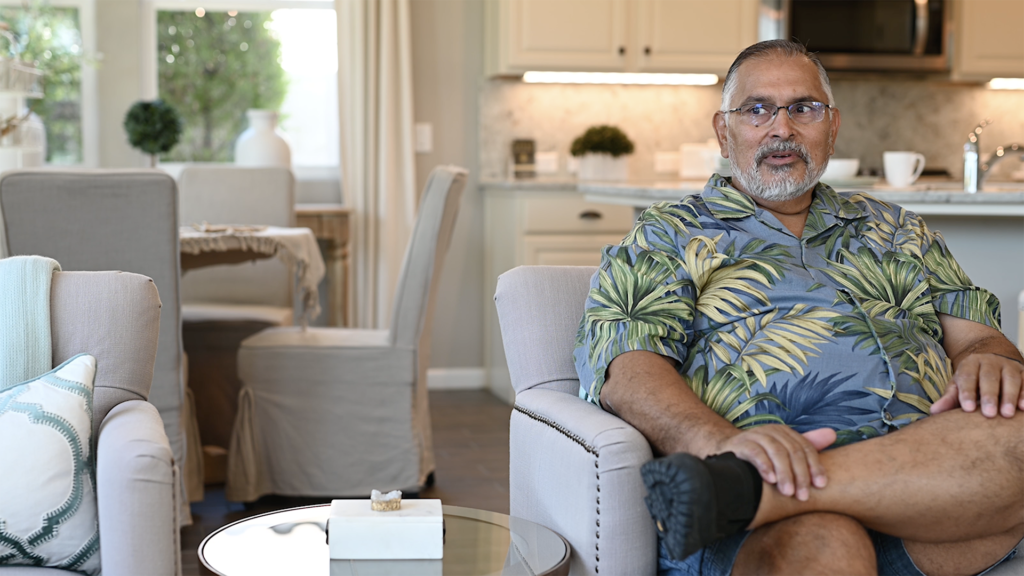 A middle-aged man with a beard and glasses, wearing a colorful tropical shirt, sits relaxed on a couch in a well-lit, modern living room with decor in soft tones.
