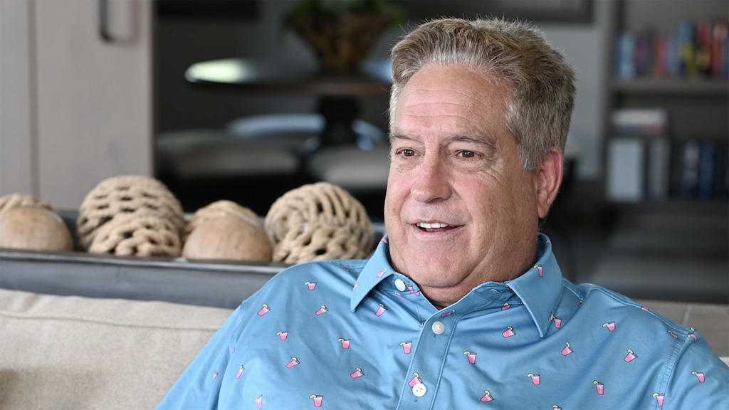 A smiling middle-aged man in a blue polo shirt with pink flamingo prints sits on a beige sofa in a well-lit living room with bookshelves in the background.