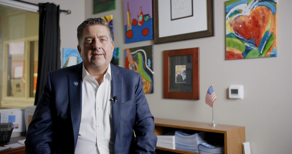 A professional man in a suit sits at a desk in an office decorated with colorful paintings and a small american flag, looking directly at the camera with a slight smile.