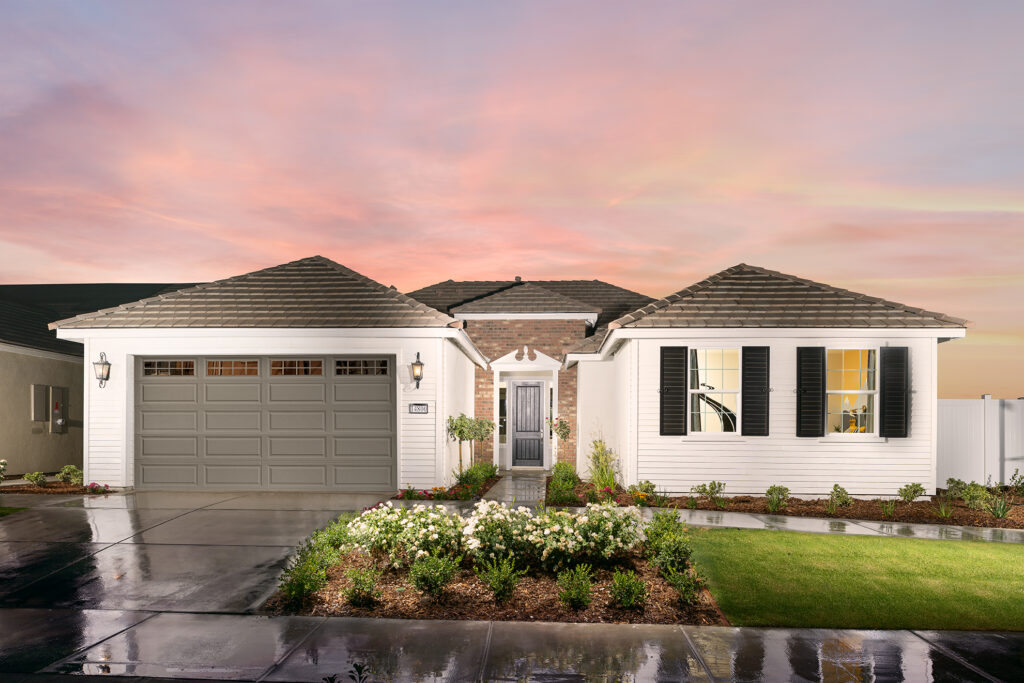 A modern single-story house with a gray facade, large garage door, and white-trimmed windows, surrounded by a manicured lawn and flowering bushes. a vibrant sunset sky looms in the background.