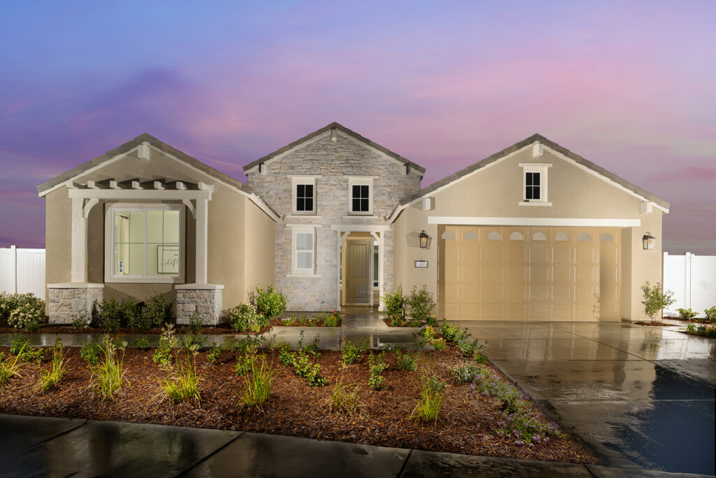 A modern single-story house with a stone facade and wet driveway under a vibrant sunset sky, surrounded by a landscaped front yard and white fence.