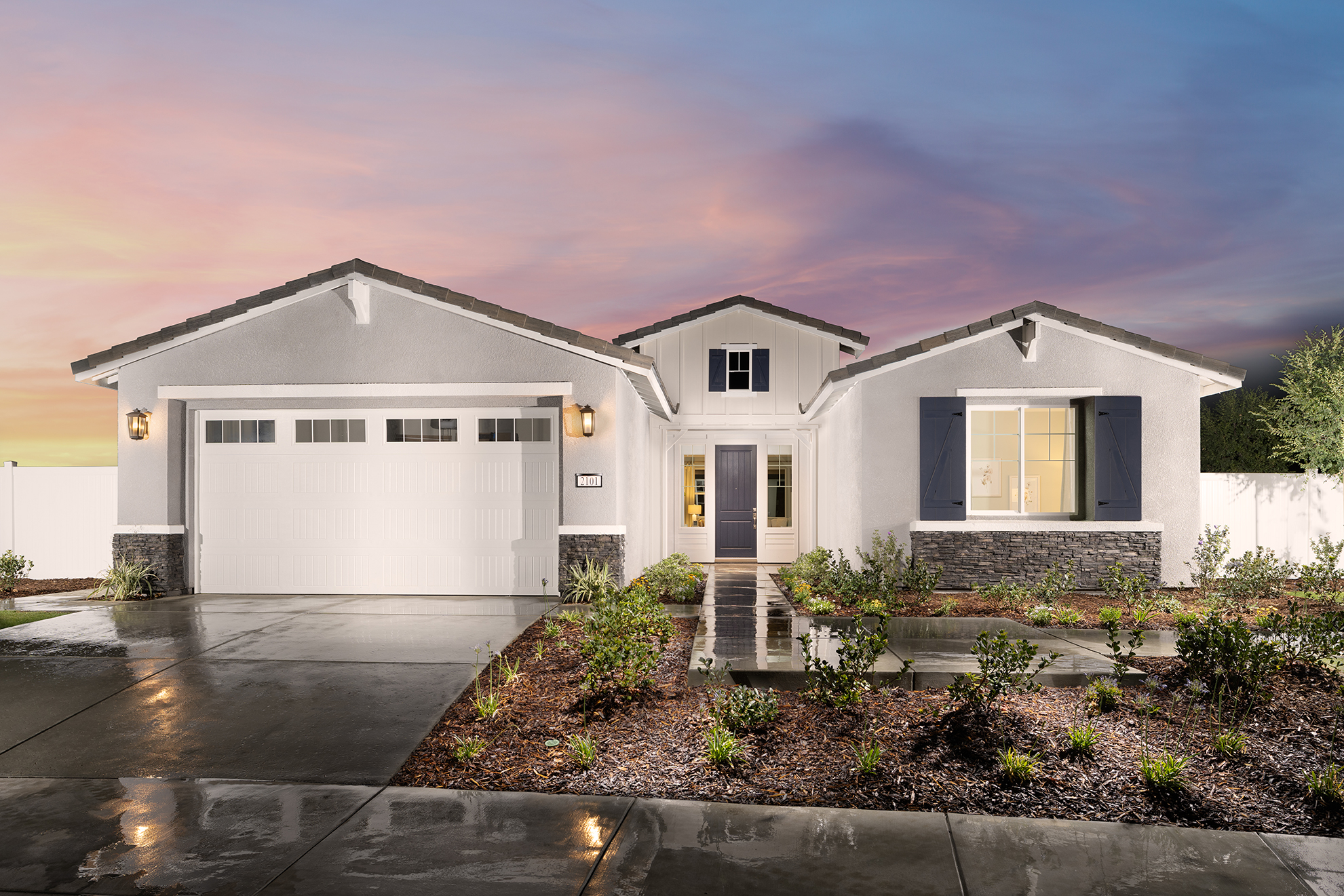 A modern single-story house with a white exterior and a two-car garage under a sunset sky. the front yard is landscaped with young trees and shrubs.