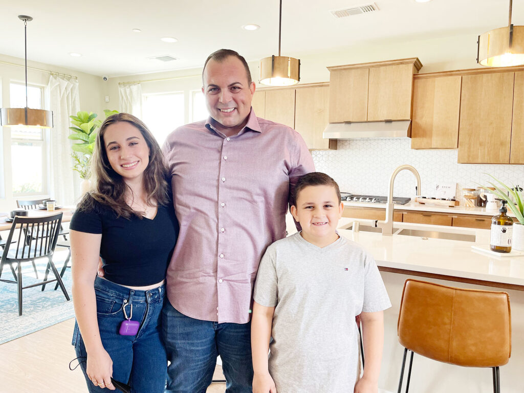 A family of three smiling in a bright kitchen with modern decor, featuring wooden cabinets, a woman, a man, and a young boy huddled together happily.