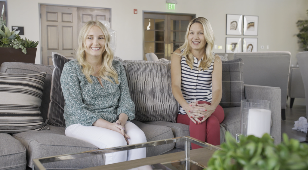Two women sitting and smiling in a living room with modern decor. one wears a green blouse and white pants, while the other sports a striped top and red pants.