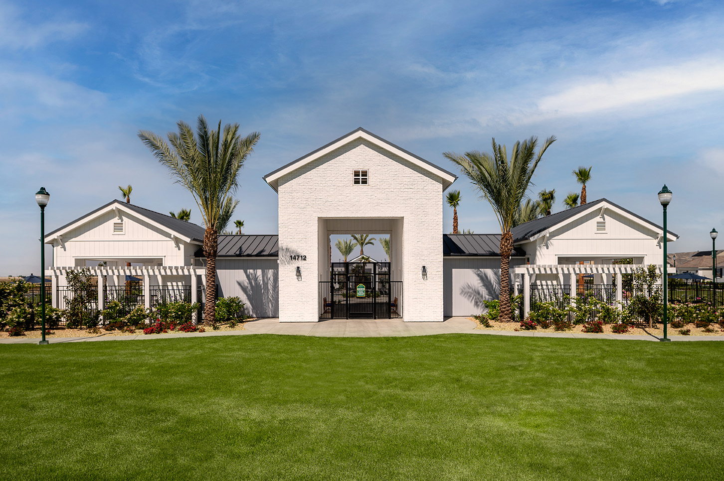 A white modern building with a symmetrical design, featuring a central archway and flanked by rows of palm trees, situated on a well-manicured lawn under a clear sky.