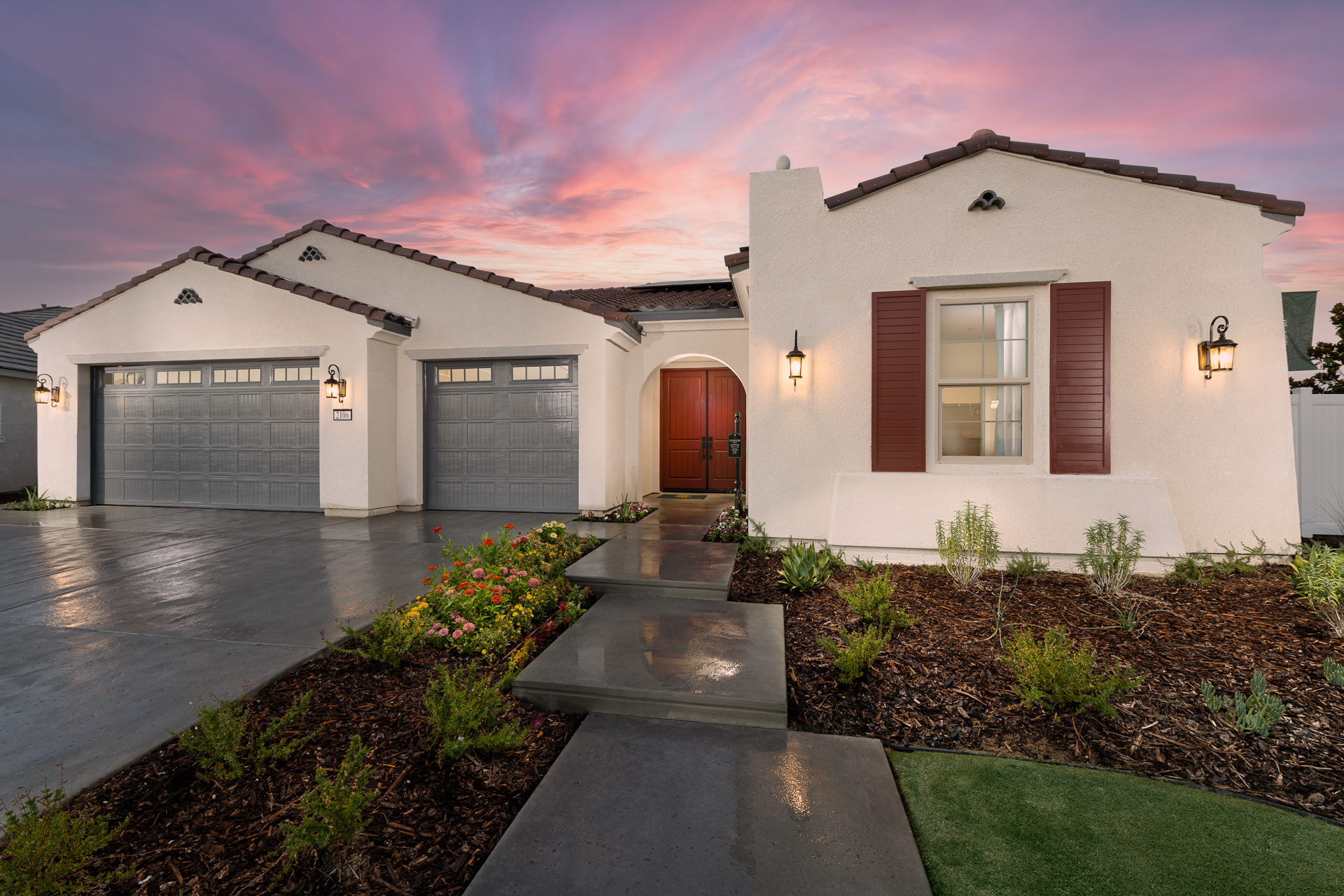 A modern suburban home with white stucco walls and gray roofs, featuring a double garage and a landscaped pathway leading to a wooden front door, set against a vibrant sunset sky.