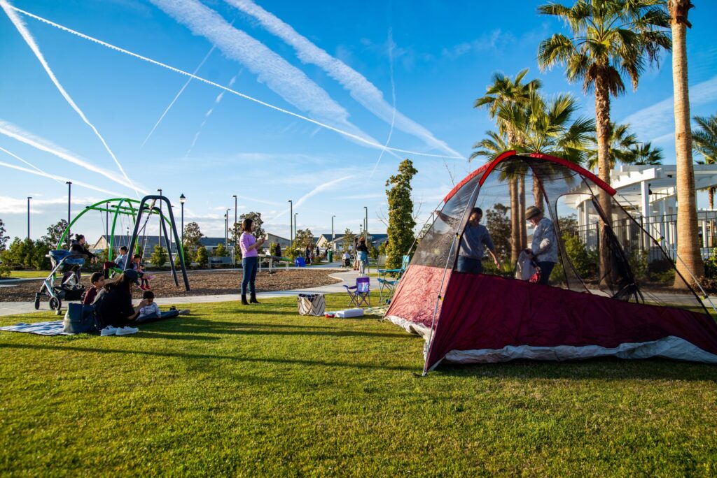 A park scene with families enjoying a sunny day; children play on swings and a tent is set up on the grass, surrounded by palm trees and a clear sky with airplane trails.