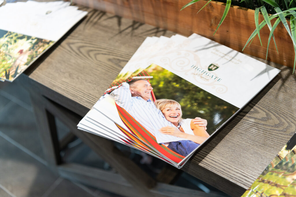 Brochures featuring a joyful elderly couple in a hammock are displayed on a wooden table, with a potted plant nearby, promoting highgate senior living.