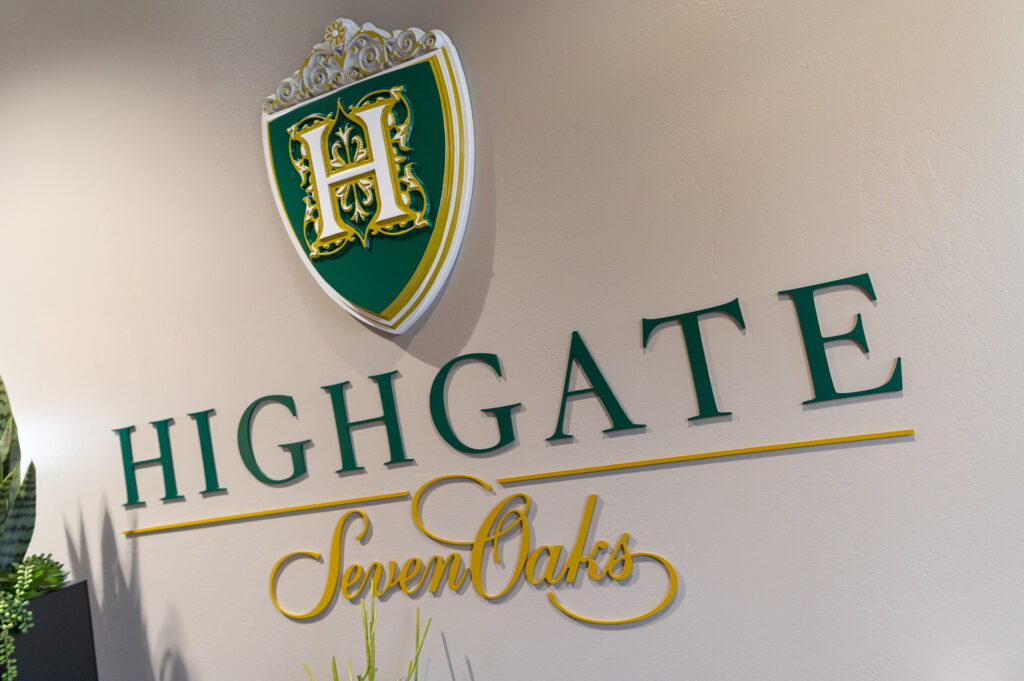 An elegant logo mounted on a wall, featuring a shield with a golden letter "h" and green details, above the gold script words "highgate" and below it "seven arts.