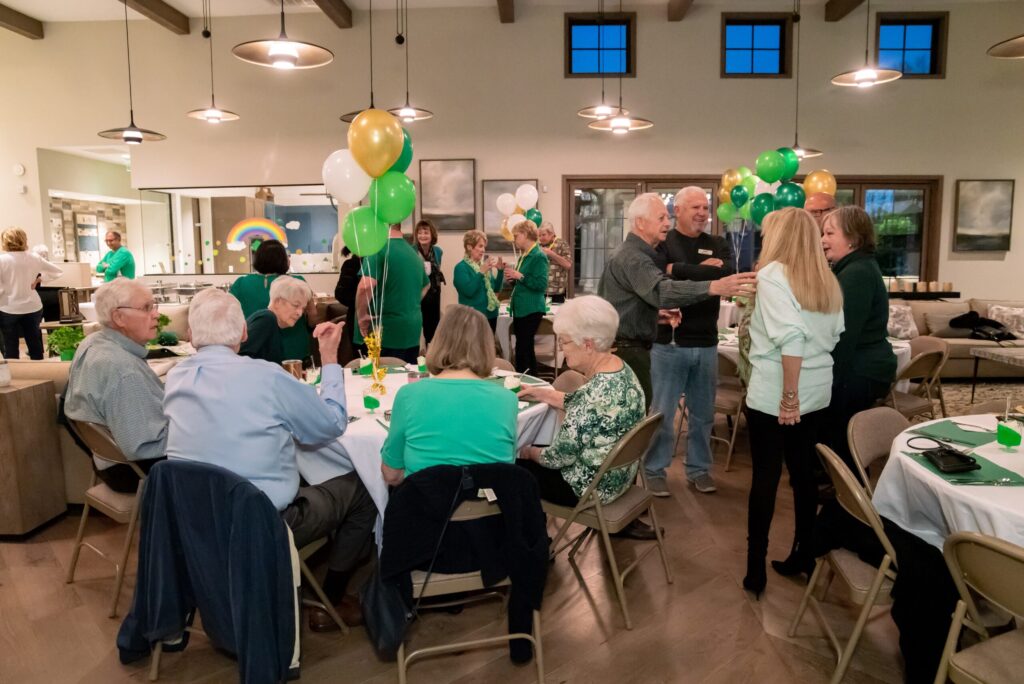 A lively senior citizen gathering in a spacious room decorated with green and yellow balloons. people are chatting and enjoying meals at tables, while two people greet each other with a handshake in the center.