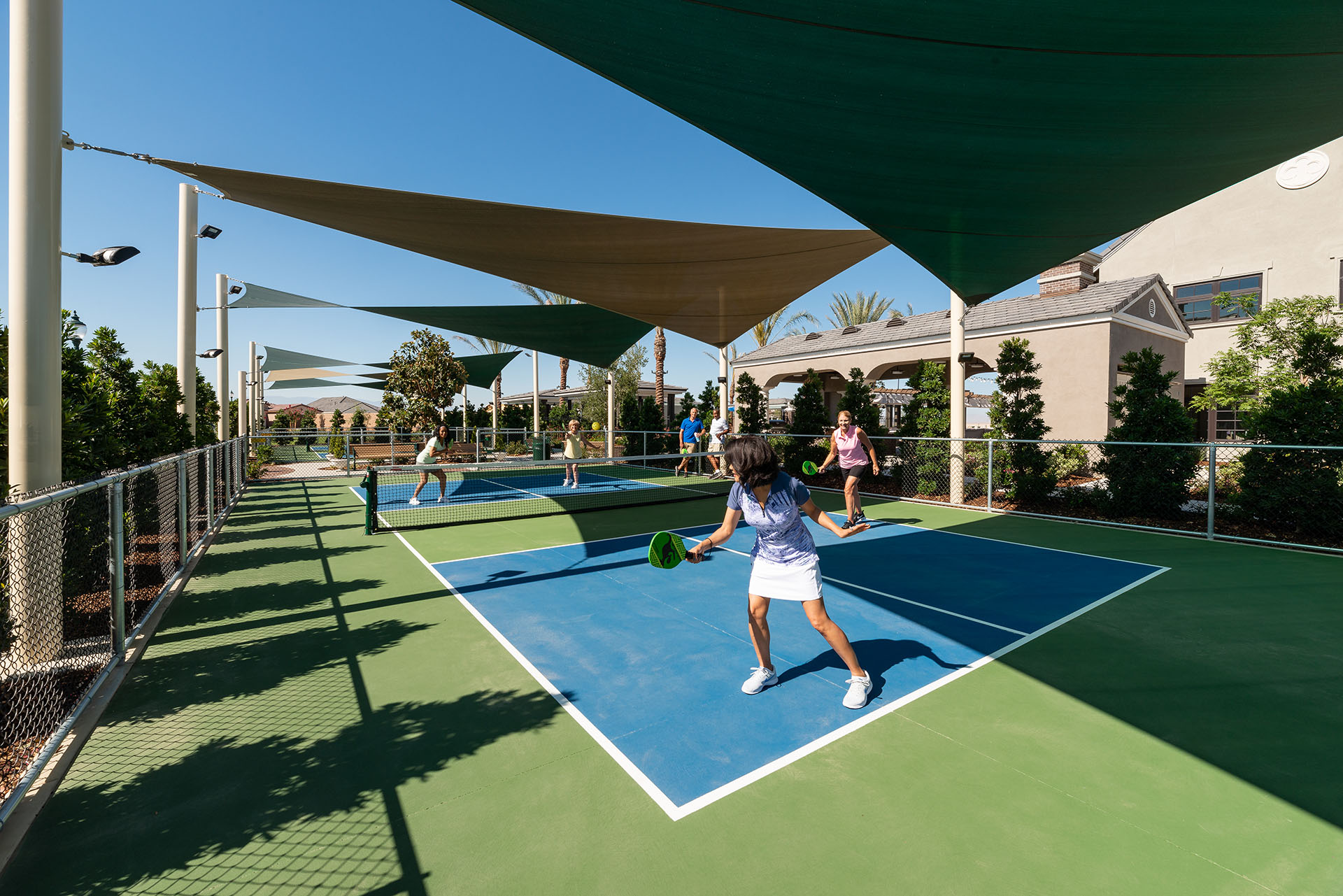 A sunny outdoor pickleball court with players engaged in a game under protective sunshades. residential buildings are visible in the background.