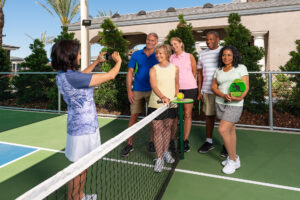 A diverse group of six older adults, three women and three men, pose smiling on a pickleball court. one woman is capturing the photo while others, in athletic wear, hold paddles or pickleballs. greenery and a clubhouse in the background.