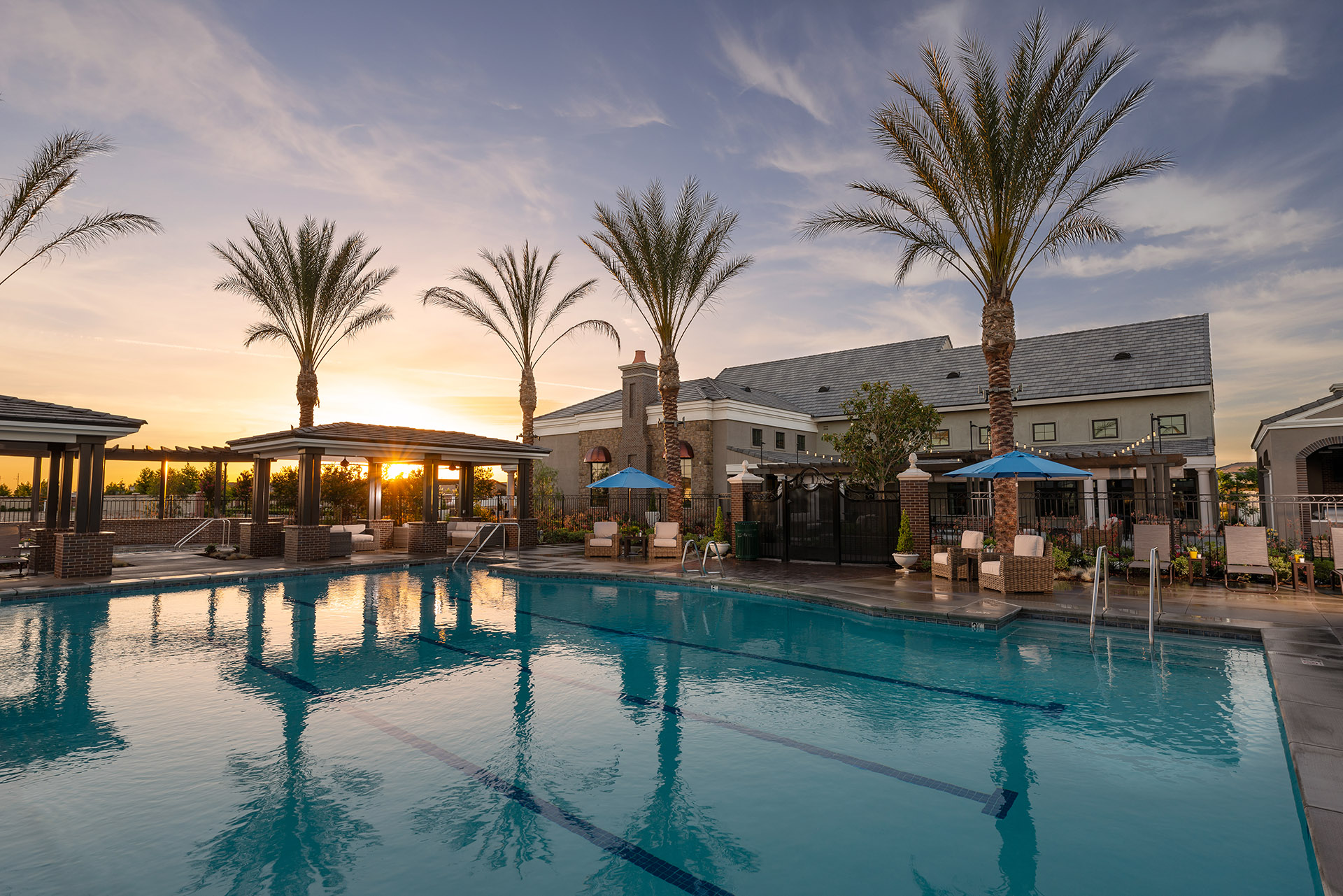 A serene poolside view at sunset with calm waters reflecting palm trees, under a clear sky, adjacent to a cozy building and lounging areas.