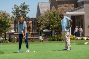 A woman in a striped shirt prepares to putt on a mini golf course under the sun, watched attentively by a man in a white polo shirt. the course is lush green with other players in the background.