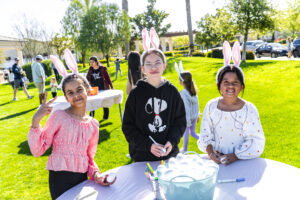 Three children wearing bunny ears stand around a table at a sunny outdoor event, smiling at the camera. one of them is decorating an egg.