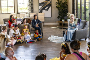 A storyteller in costume reads to a group of children and adults in a modern lounge area. the children, some dressed in costumes, listen attentively, surrounded by light decor and large windows.