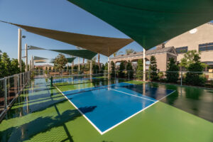 A pickleball court with a bright blue playing area, surrounded by green areas, is shaded by large triangular fabric canopies. The court is enclosed by a chain-link fence, with trees and a building visible in the background. The sky is clear and sunny.