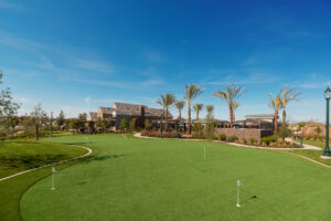 A putting green with several flagged holes in a well-maintained grassy area. The background features a clubhouse with a patio, surrounded by palm trees and landscaping, under a clear blue sky. A lamp post stands to the right.