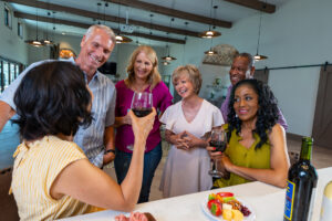 A diverse group of six mature adults gather around a table, smiling and chatting. Two women are holding glasses of red wine. The table has a cheese and fruit plate. The setting seems to be a cozy indoor space with modern decor and pendant lights.