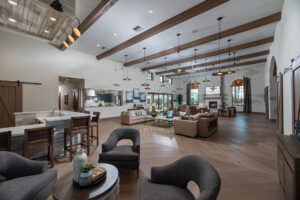 A spacious, modern living area featuring a large gray sectional sofa, armchairs, and a central coffee table. The room has a high ceiling with exposed wooden beams, multiple pendant lights, and a fireplace. Adjacent to the living space, there’s a kitchen with bar stools.